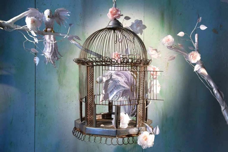 Ein dekorativer Vogelkäfig mit weißen Vögeln und Blumen aus Papier vor einem sanften Hintergrund.