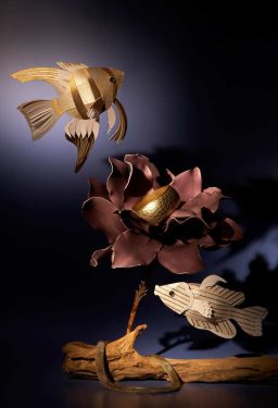 Drei goldene Fische aus Papier schwimmen über eine blühende Blume auf einem Holzstück.